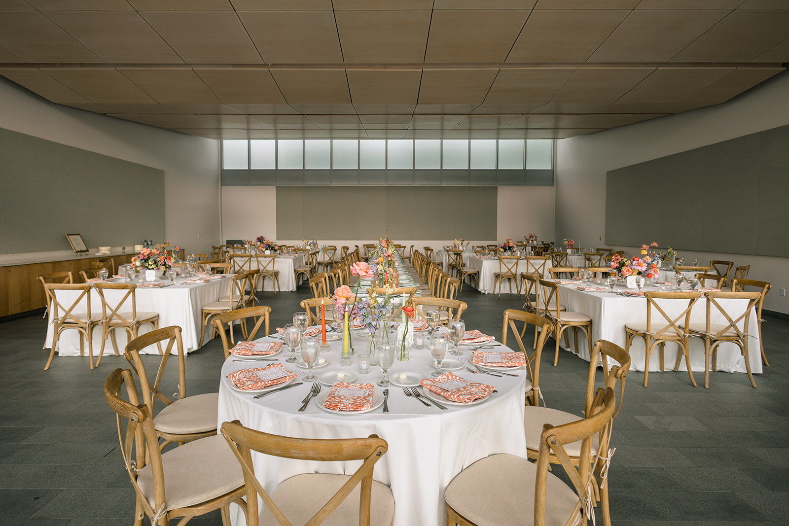 A long, elegantly set dining table is decorated with colorful flowers in vases, glassware, and plates, situated in a bright room with large windows at a garden wedding venue