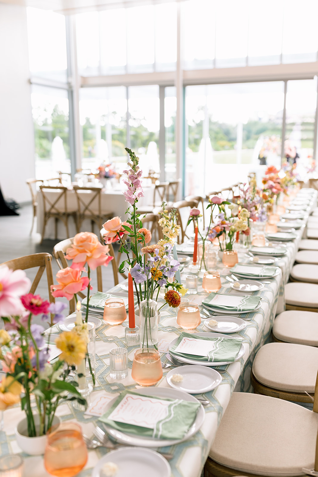 A long, elegantly set dining table is decorated with colorful flowers in vases, glassware, and plates, situated in a bright room with large windows at a garden wedding venue