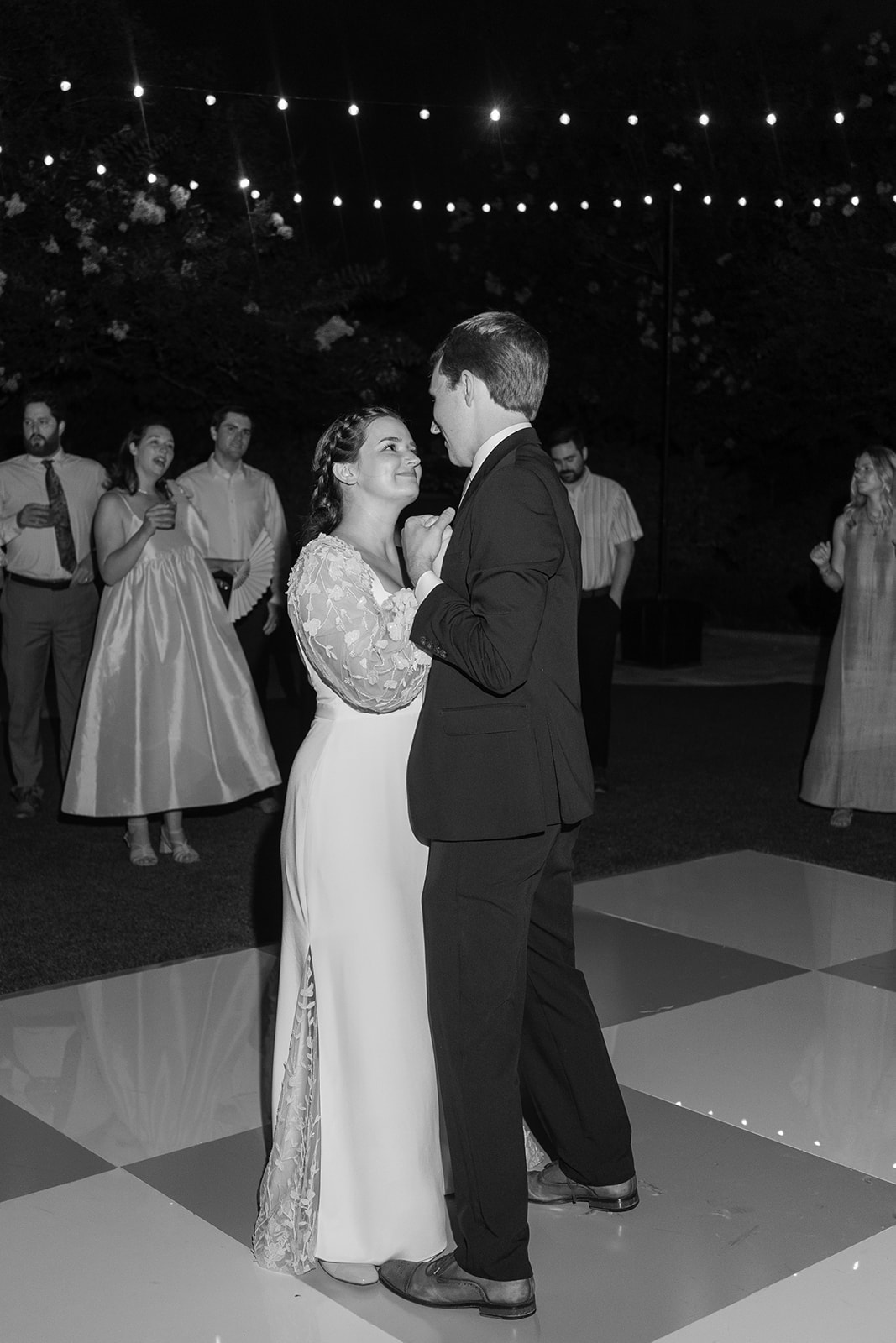 A bride and groom dance together on an outdoor checkered dance floor at night, surrounded by guests watching under string lights.