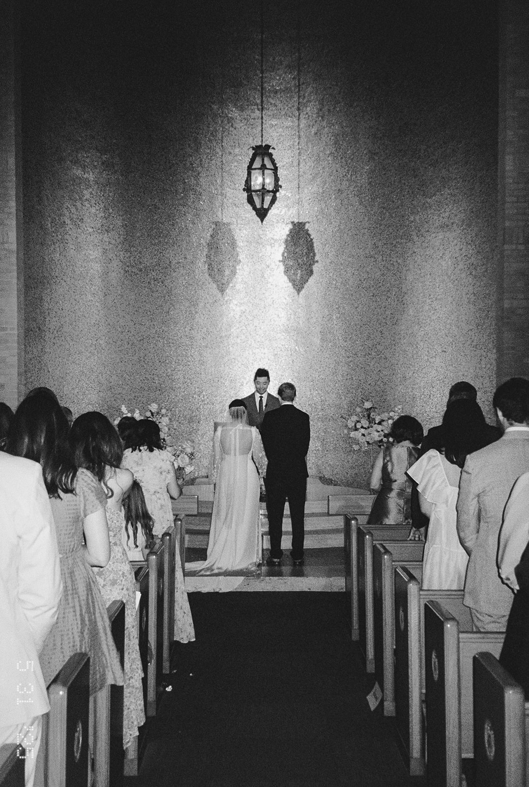 A bride and groom stand at the altar with an officiant in a church filled with golden light, as guests watch the wedding ceremony from the pews.