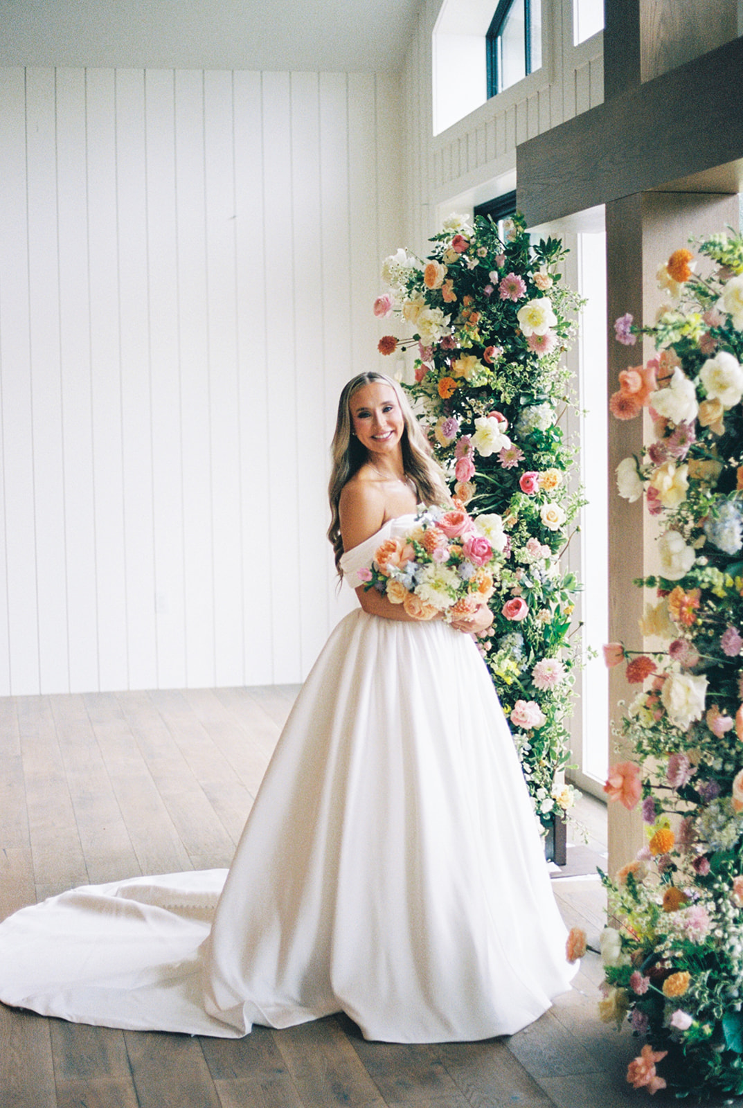 Bride in a white gown and groom in a blue suit pose outdoors, smiling, with greenery and trees in the background at the blue magnolia