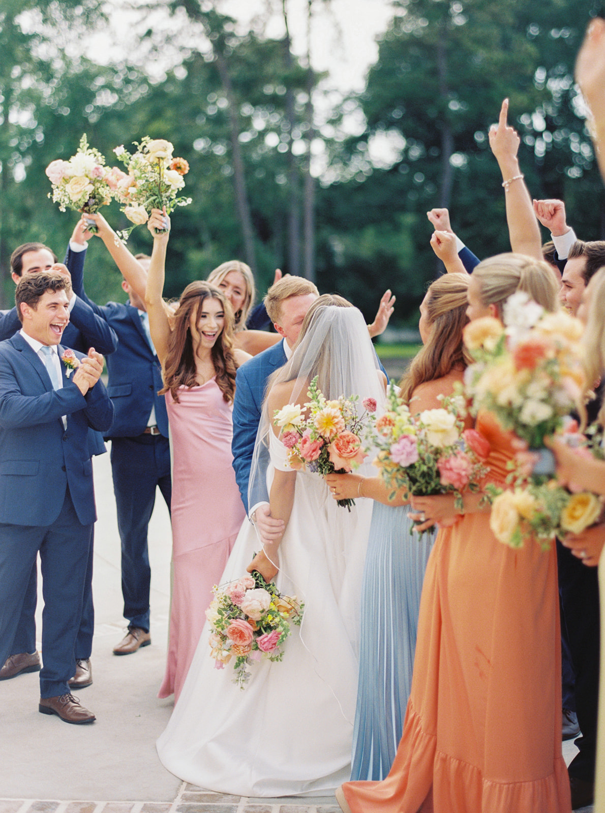 A large wedding party stands outdoors in a line, with men in dark suits and women in colorful dresses, holding bouquets, trees and grass in the background  at the blue magnolia