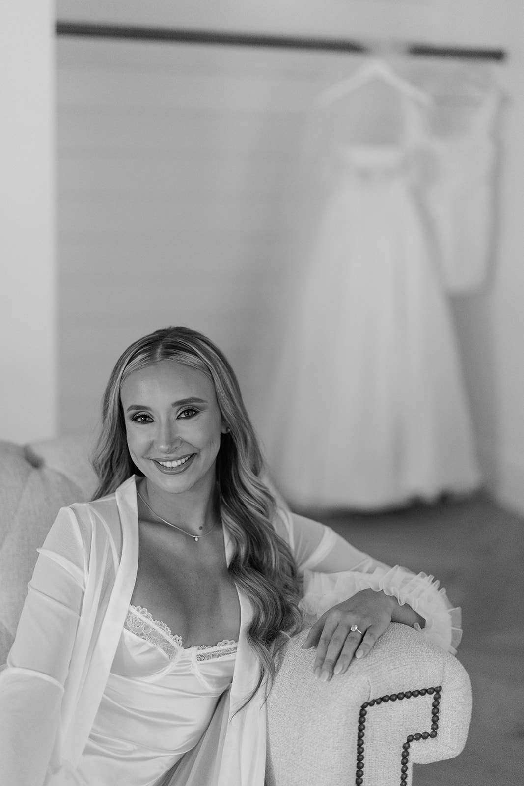 A woman in an off-the-shoulder white wedding dress stands indoors near a wall mirror and glass table, looking at the camera.