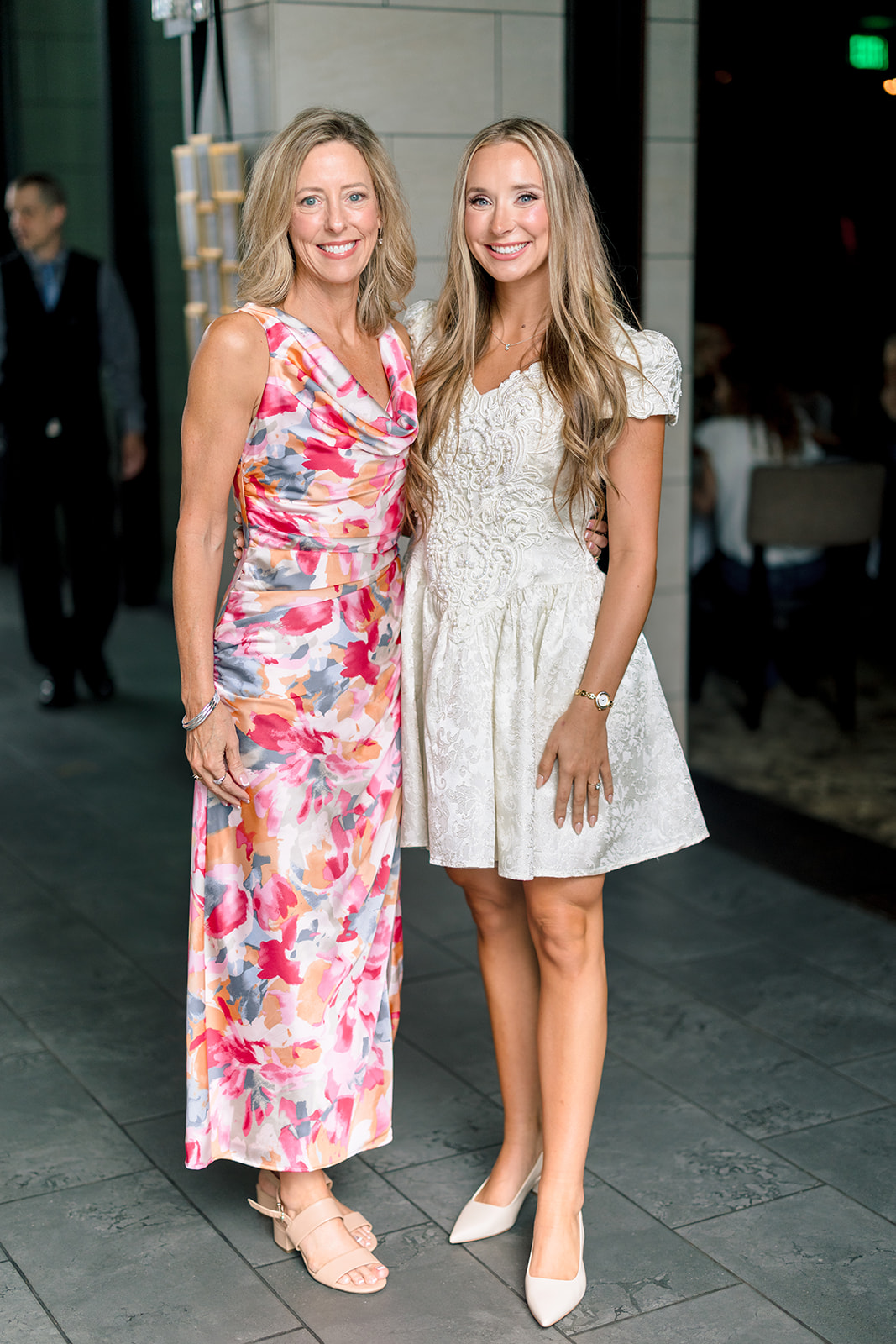 Two women stand side by side indoors; one wears a floral sleeveless dress, the other a short-sleeved white dress. Both are smiling and looking at the camera.