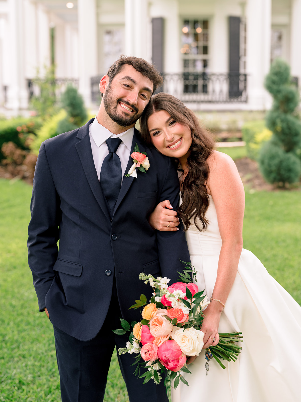A bride in a white dress holds a bouquet and stands beside a groom in a navy suit on a grassy lawn in front of a white building.