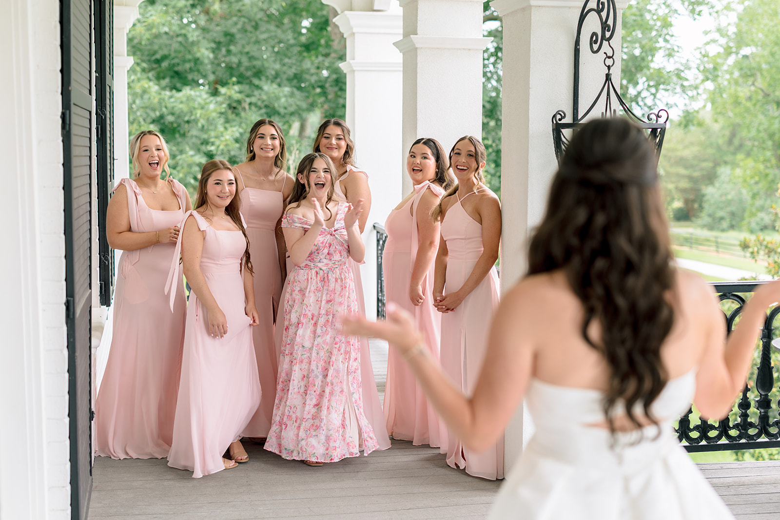 A bride in a white dress stands facing a group of bridesmaids wearing light pink dresses on a porch, with greenery visible in the background for a sandlewood manor wedding 