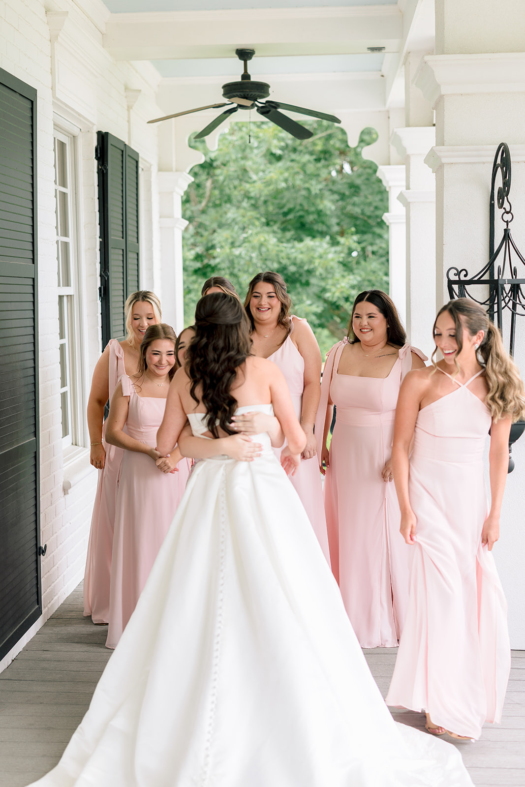 A bride in a white dress stands facing a group of bridesmaids wearing light pink dresses on a porch, with greenery visible in the background for a sandlewood manor wedding 
