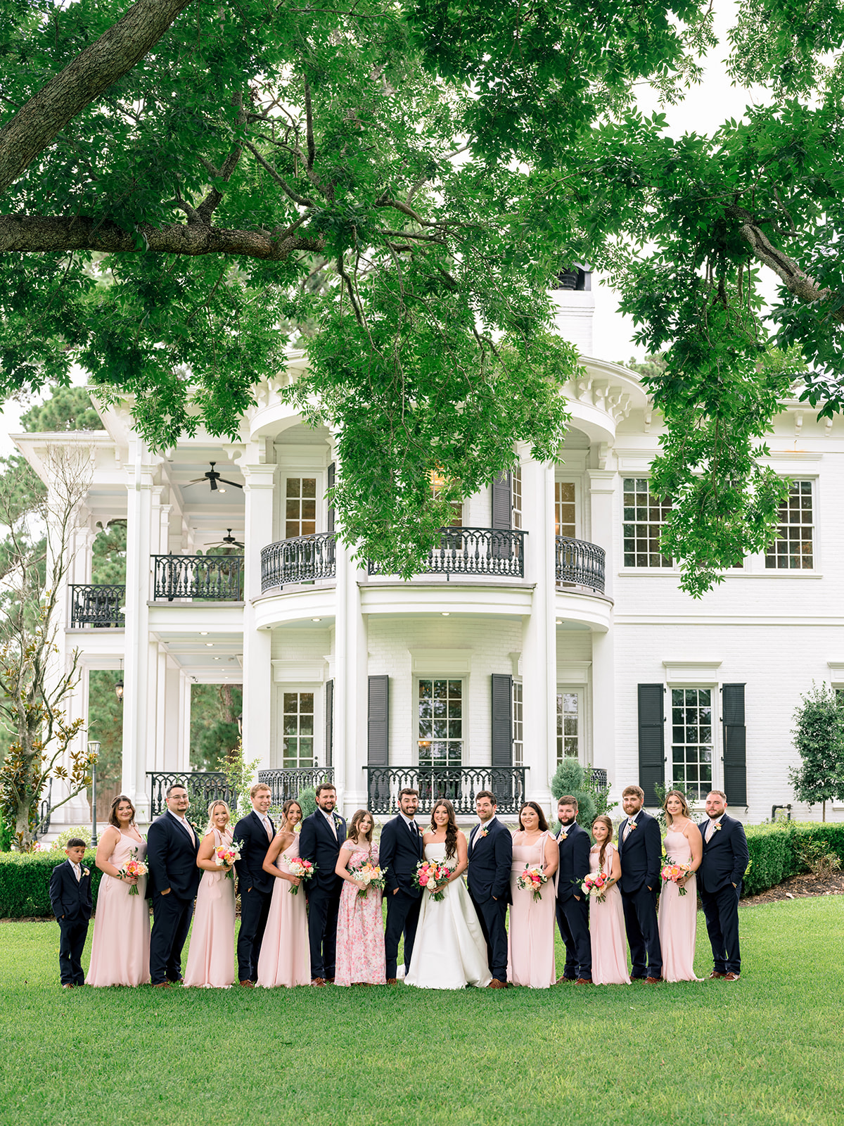 A bride and groom stand outside with bridesmaids in pink dresses and groomsmen in navy suits, all holding bouquets, in front of a white building for a Sandlewood Manor wedding