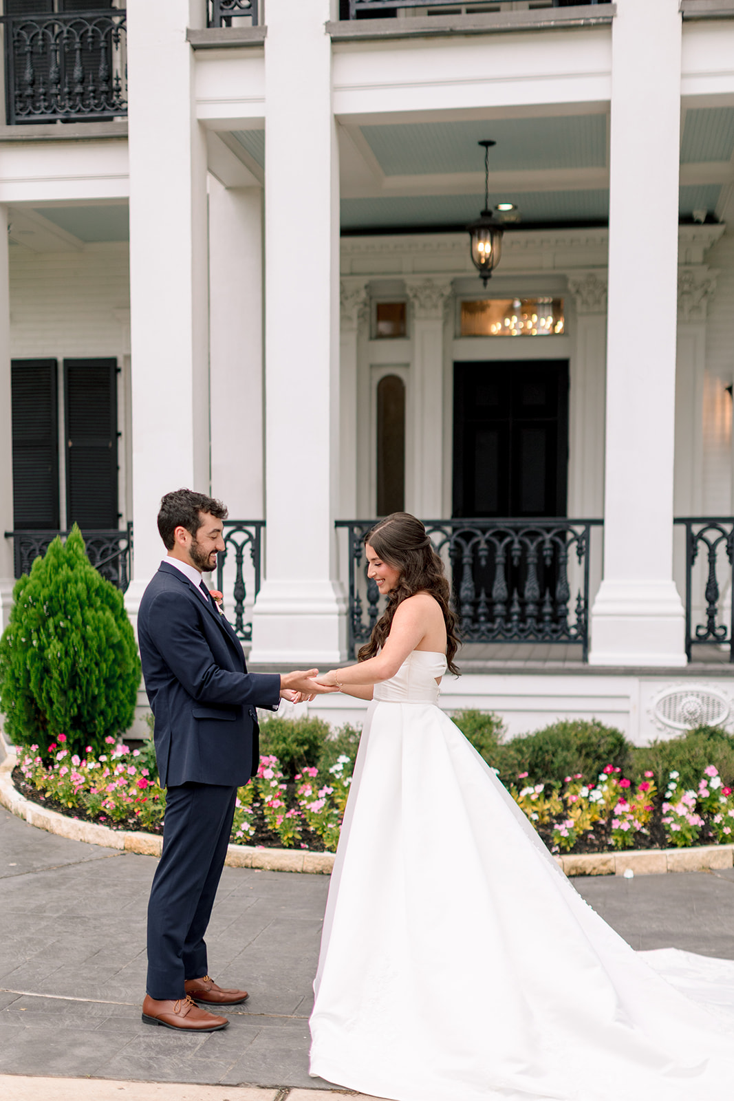 Bride and groom stand facing each other outside near a white fountain, exchanging vows or letters on their wedding day.