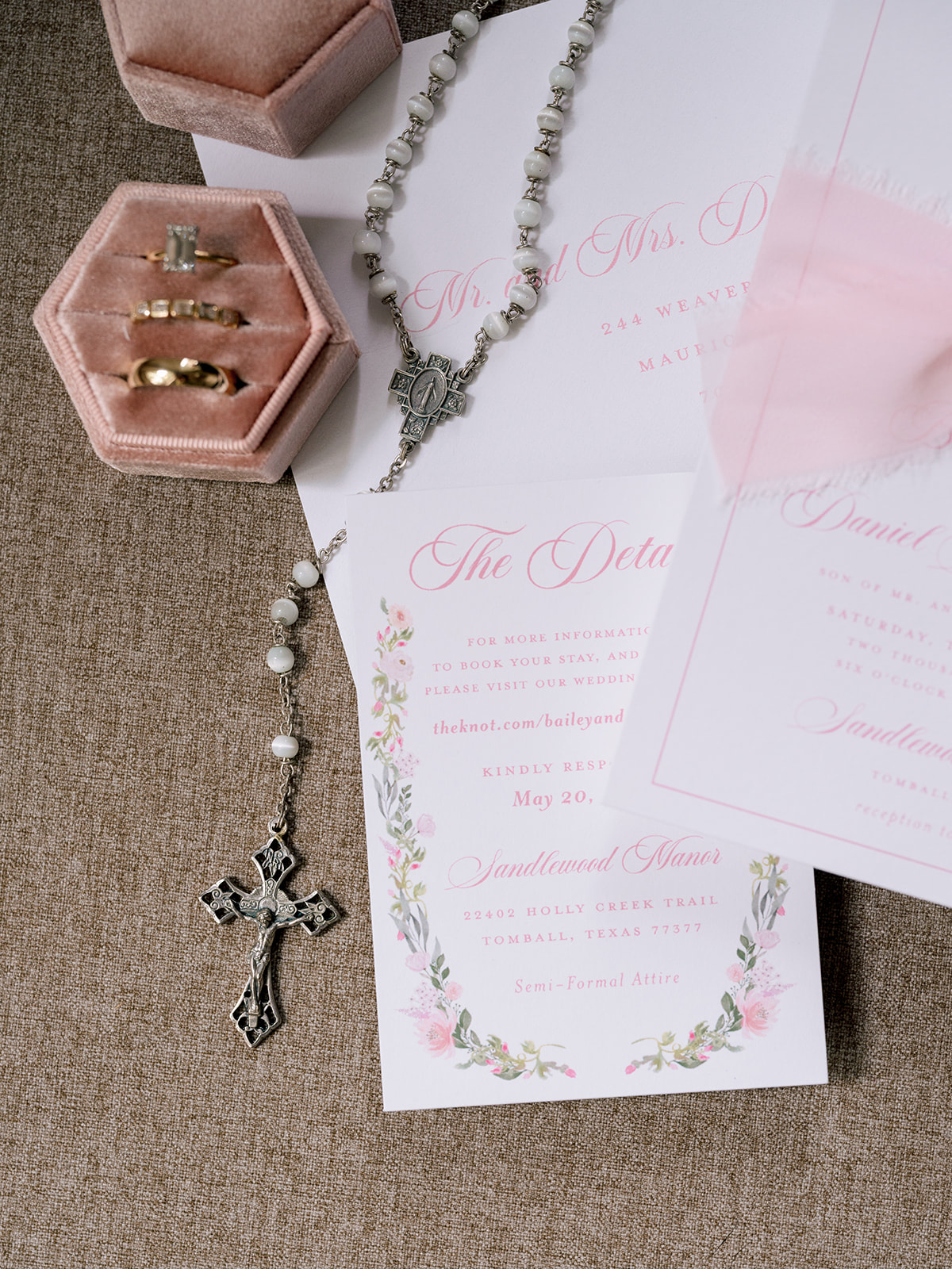 Wedding invitation with floral border, two gold rings in a pink ring box, and a rosary with a silver cross arranged on a brown fabric surface.