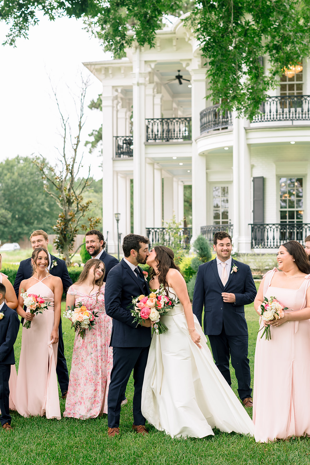 A bride and groom stand outside with bridesmaids in pink dresses and groomsmen in navy suits, all holding bouquets, in front of a white building for a Sandlewood Manor wedding
