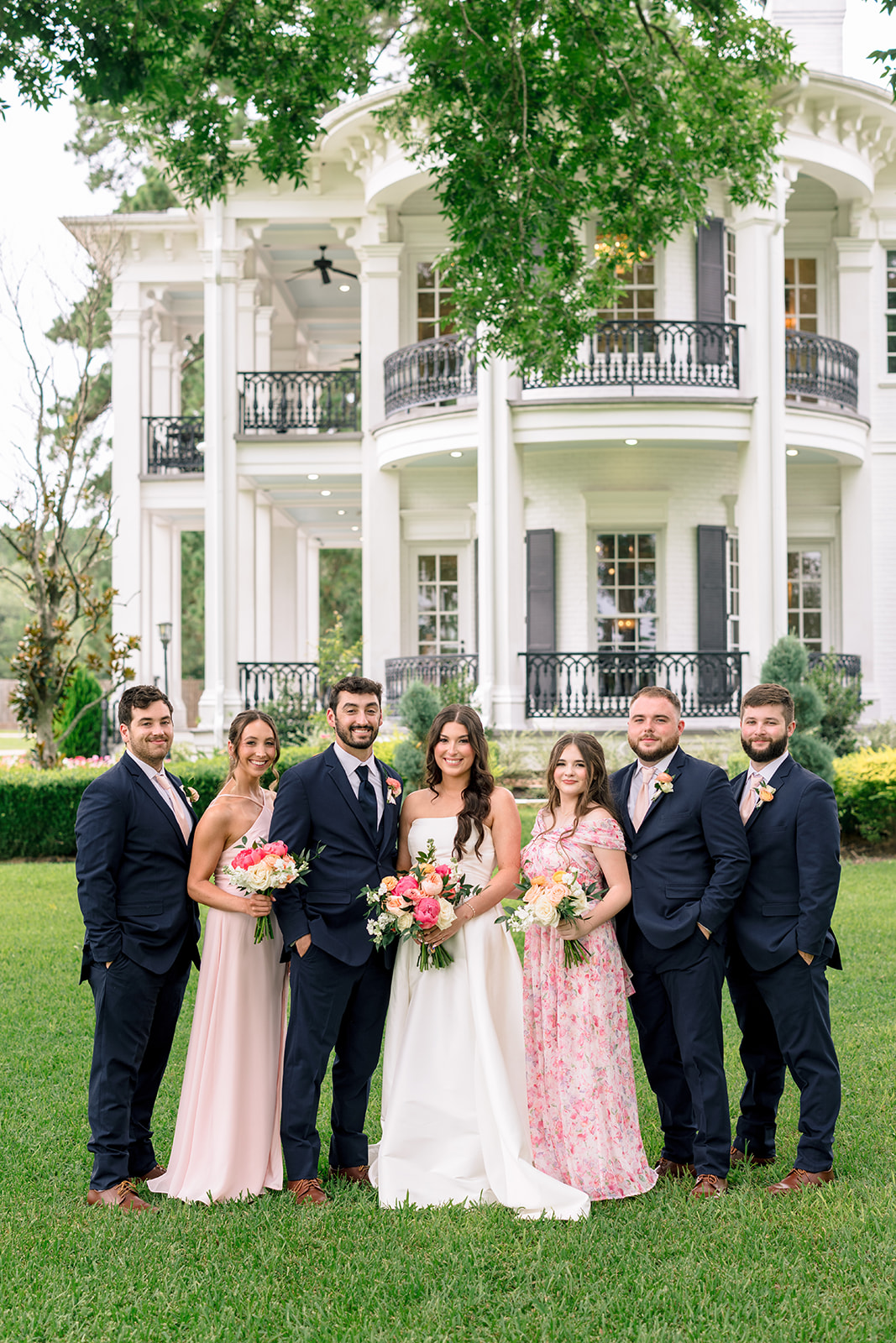 A bride and groom stand outside with bridesmaids in pink dresses and groomsmen in navy suits, all holding bouquets, in front of a white building for a Sandlewood Manor wedding