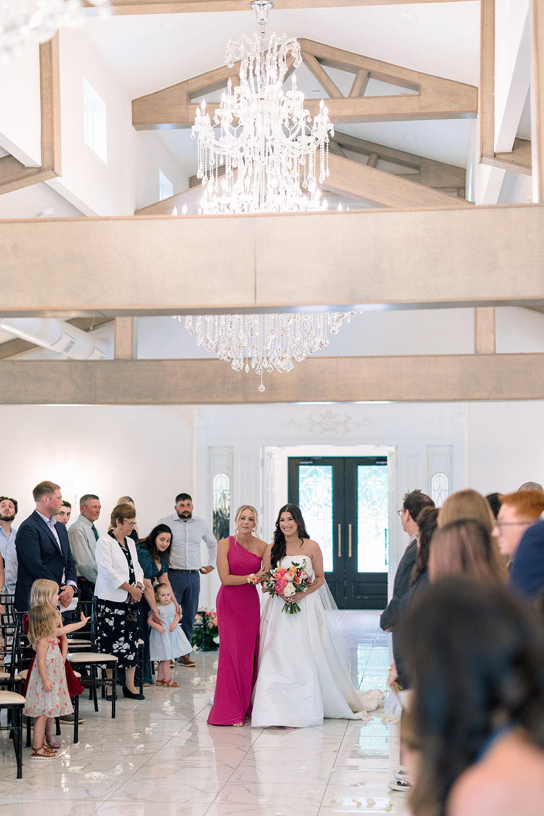 A bride walks down the aisle towards the groom during a wedding ceremony in a bright, indoor venue, with guests seated and standing on either side.
