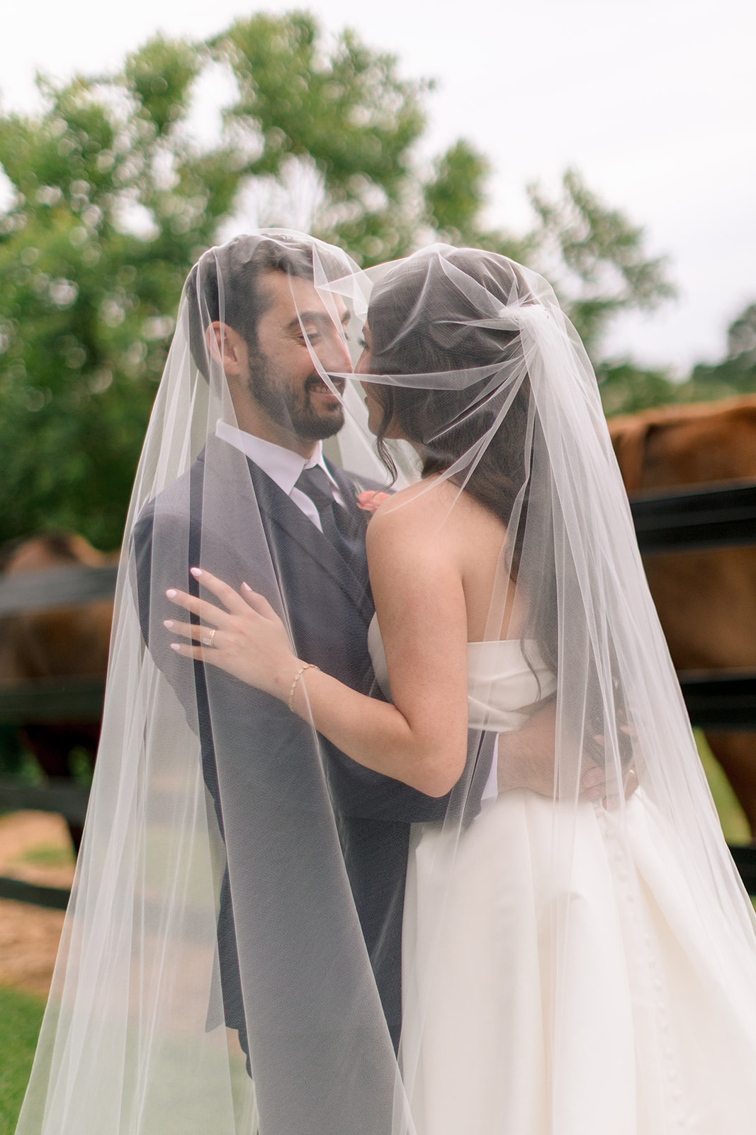 A bride in a white dress holds a bouquet and stands beside a groom in a navy suit on a grassy lawn in front of a white building.