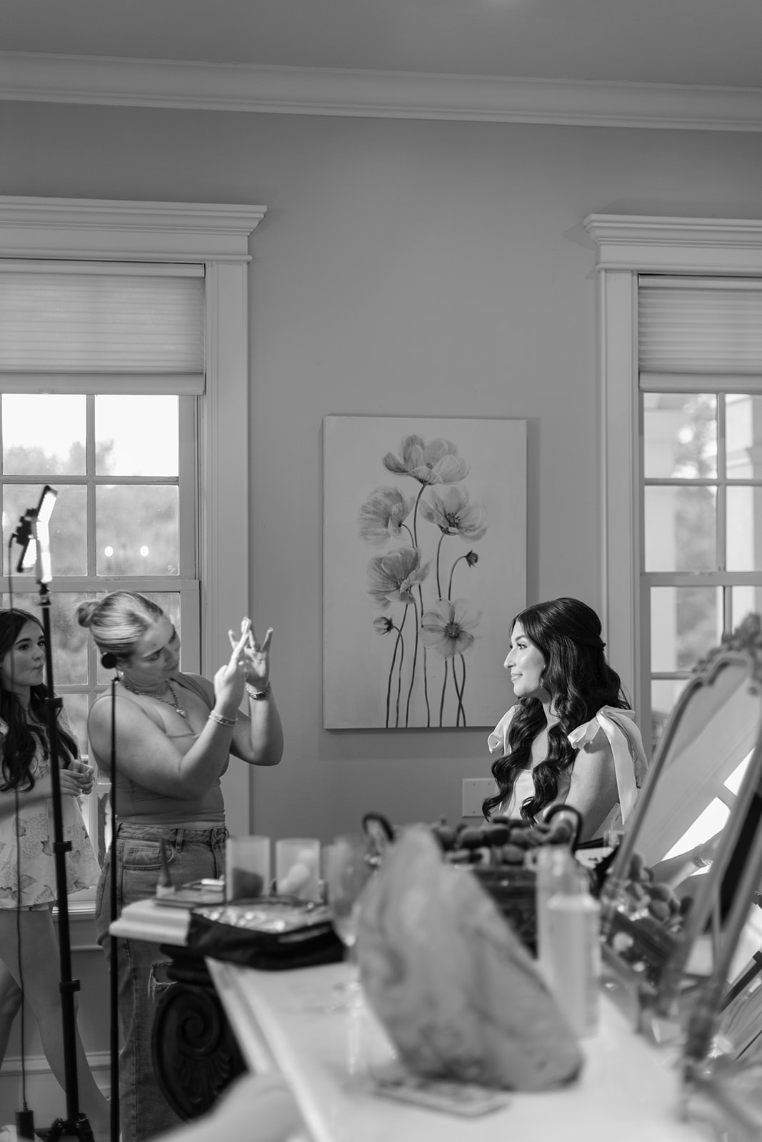 Three women are in a room with makeup and hair tools; one styles another’s hair while the third observes. A floral painting and two windows are visible in the background.