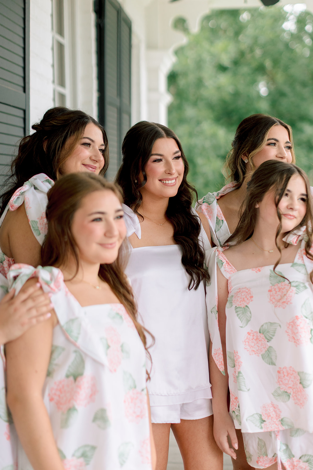 Six young women in matching floral pajamas and slippers stand together on a porch, smiling at the camera. One woman in the center wears a different white pajama set for a sandlewood manor wedding 