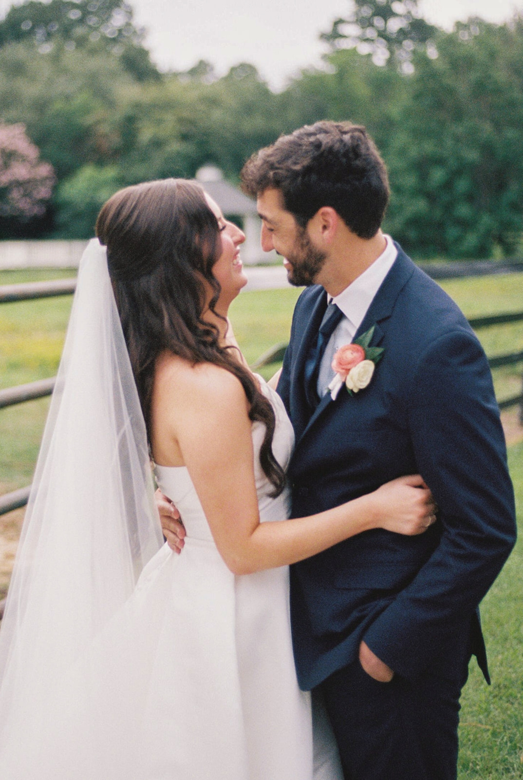 A bride in a white dress holds a bouquet and stands beside a groom in a navy suit on a grassy lawn in front of a white building.