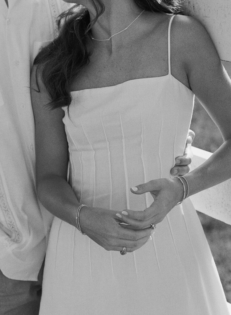 Black-and-white detail shot of Adalyn's clasped hands resting at her waist, showing her engagement ring, layered bracelets, and necklace as Miles holds her close.