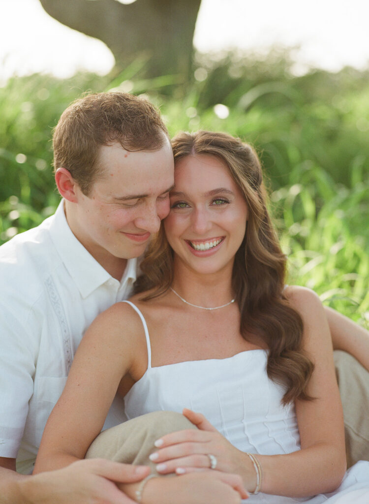 Adalyn laughs brightly at the camera while Miles nuzzles close to her forehead with his eyes closed, the two sitting together against a backdrop of blurred tall grass and a shadowed tree trunk.