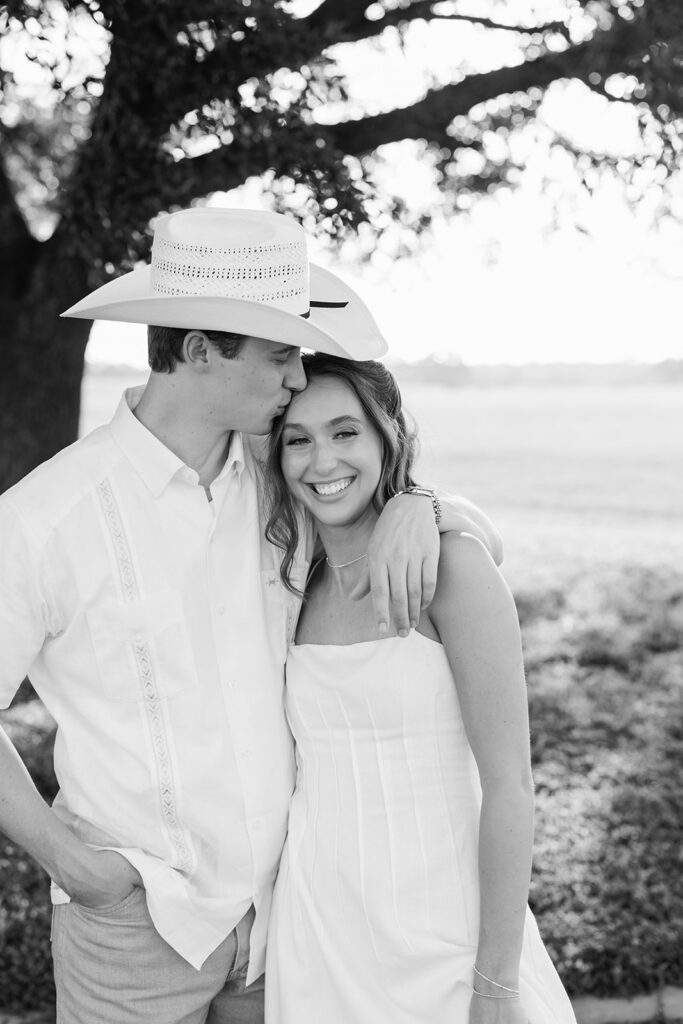 Black and white photo of Miles kissing Adalyn's forehead as she smiles with her eyes closed, his cowboy hat on and the open field blurred behind them.