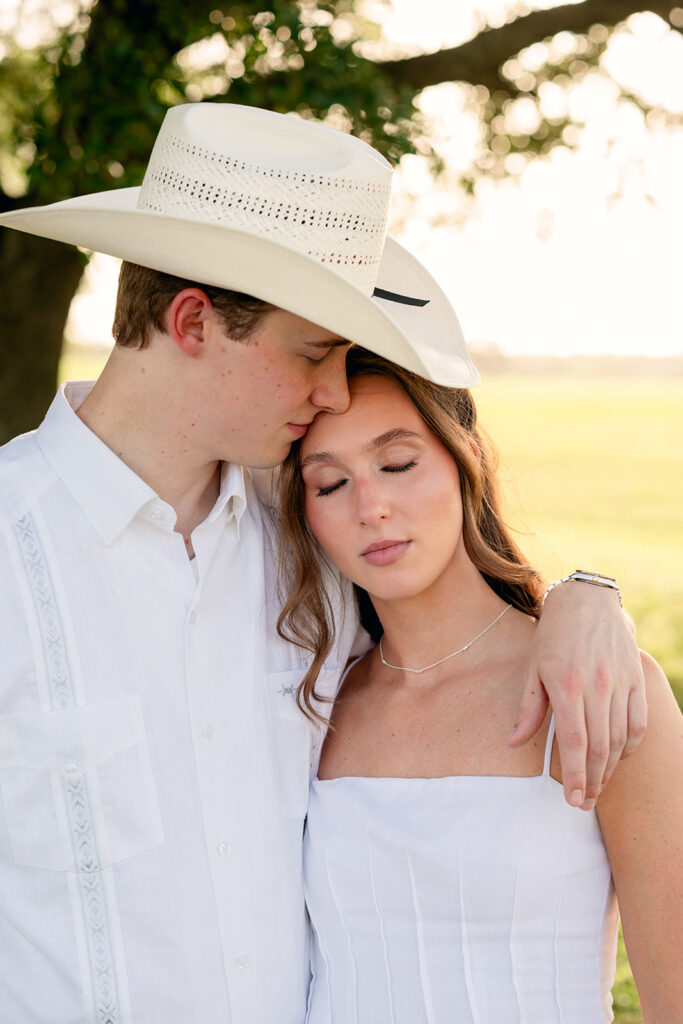 Miles, wearing his white cowboy hat, leans close to Adalyn's forehead as she closes her eyes, the open field glowing softly behind them in golden light.