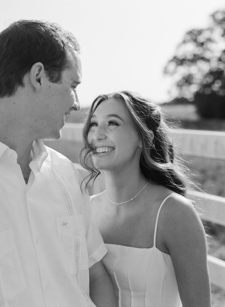 In black and white, Miles gazes down smiling at Adalyn as she laughs, the white ranch fence and an oak tree softly blurred behind them.