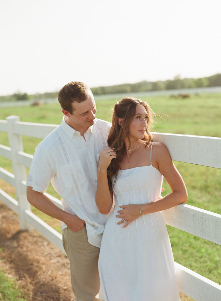 Adalyn looks off to the side with her hand in her hair as Miles gazes at her, both leaning near a white ranch fence with distant grazing animals visible in the pasture behind them.