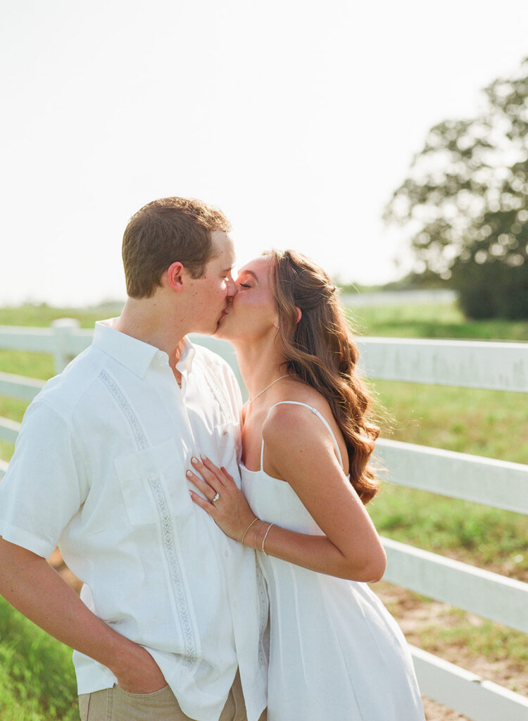 Adalyn and Miles share a kiss in profile beside a white ranch fence, a large oak tree softly blurred behind them in the open field.