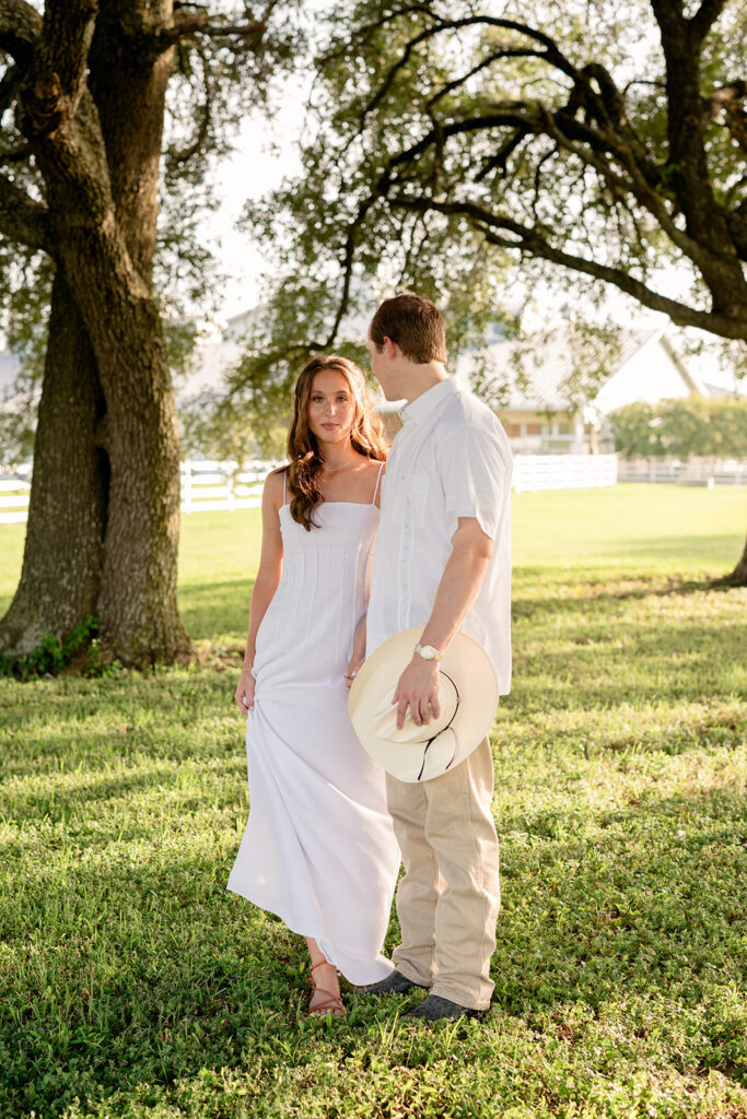 Adalyn and Miles stand together beneath a sprawling oak tree with the white ranch fence and farmhouse behind them, Miles holding his cream cowboy hat at his side.