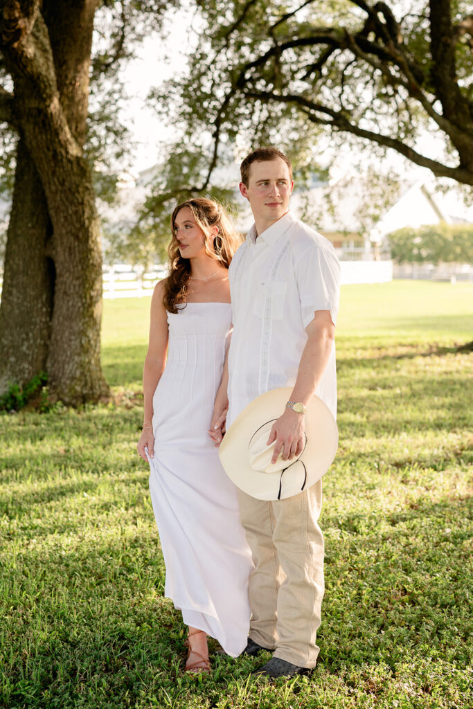 Adalyn and Miles hold hands beneath the oak tree, the white ranch fence and farmhouse softly blurred behind them as Miles carries his cowboy hat.