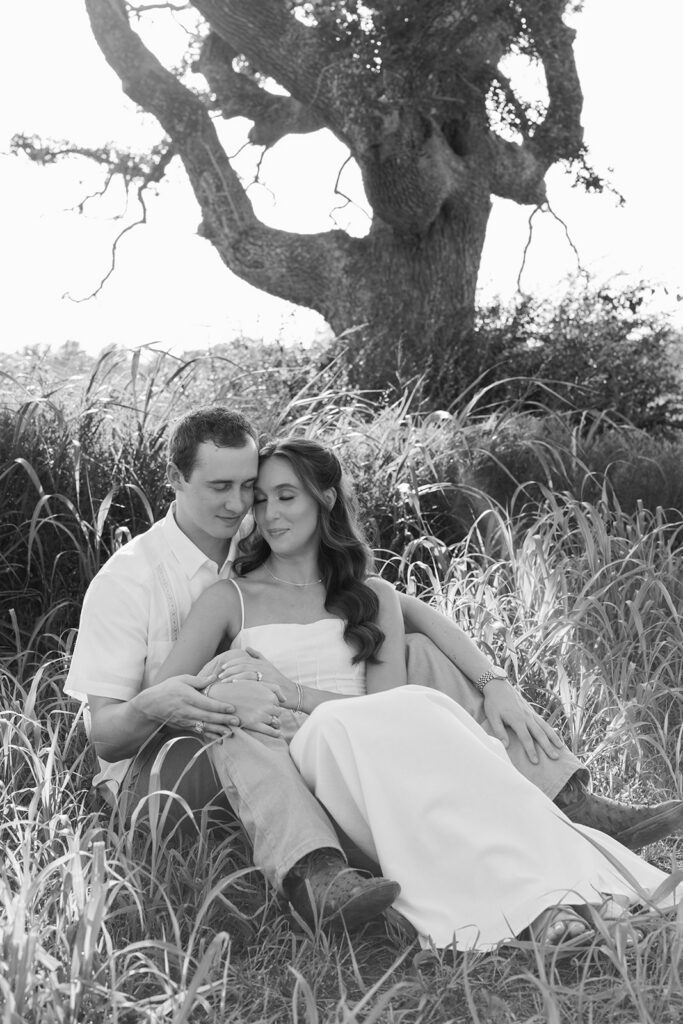 Adalyn and Miles sit together in tall grass beneath the sprawling oak tree, foreheads touching with eyes closed, in a black-and-white portrait.