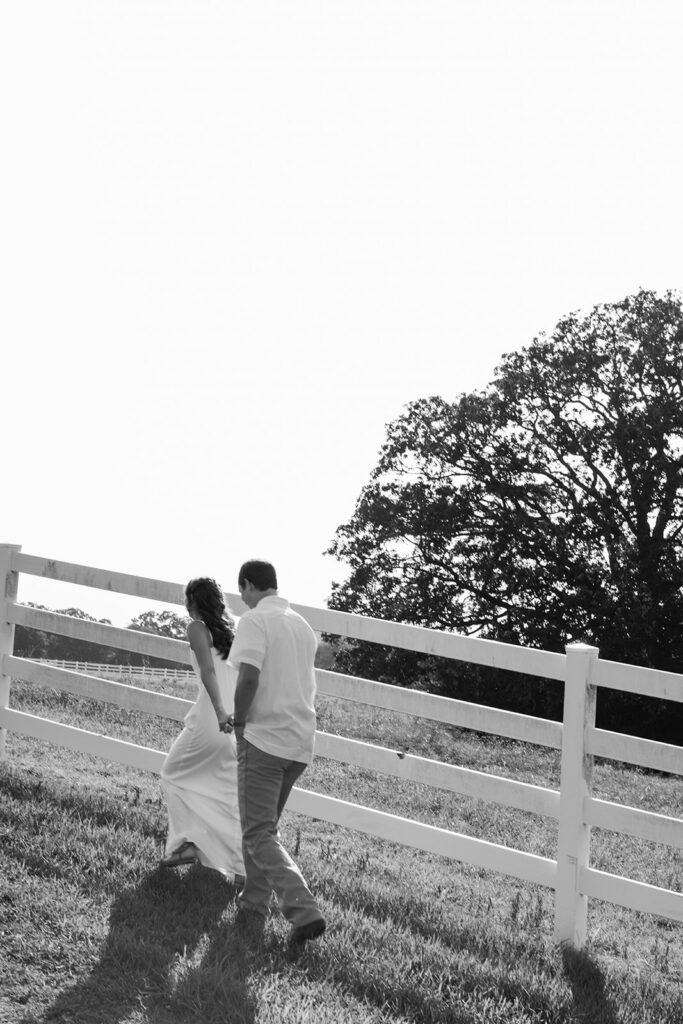 Adalyn and Miles walk hand in hand beside the ranch fence toward a large oak tree, photographed in black and white.