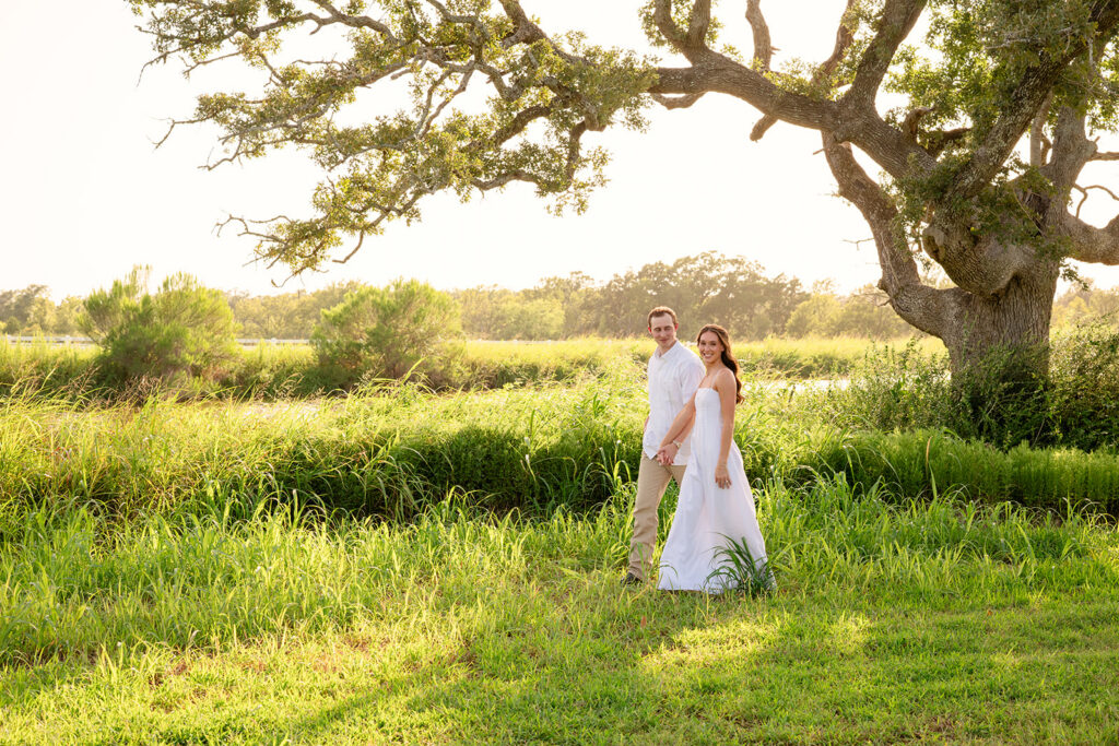 Adalyn and Miles walk hand in hand through tall grass beneath the oak tree, glancing back over their shoulders at the camera.