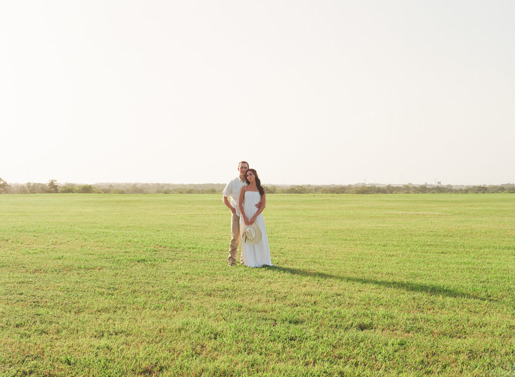 In a sunlit green pasture, Adalyn and Miles stand close together, Adalyn holding a straw cowboy hat in front of her as Miles rests his head against hers.