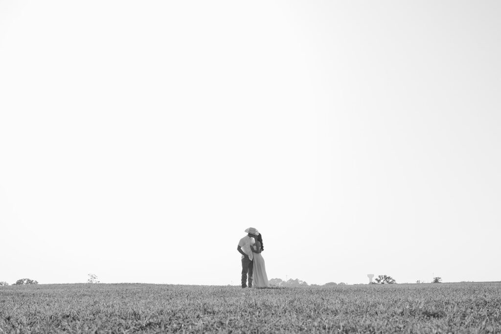 Adalyn and Miles kiss in an open Texas field, the water tower visible in the distance, captured in black and white.
