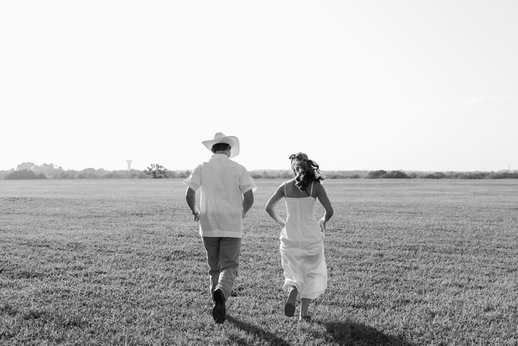 Adalyn and Miles run playfully across an open field toward the TAMU water tower on the horizon, shot in black and white.