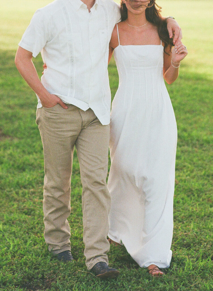 A cropped detail shot shows Miles in a white guayabera shirt and khaki pants standing beside Adalyn in her long white spaghetti-strap dress, his arm around her shoulder, against a blurred green lawn.