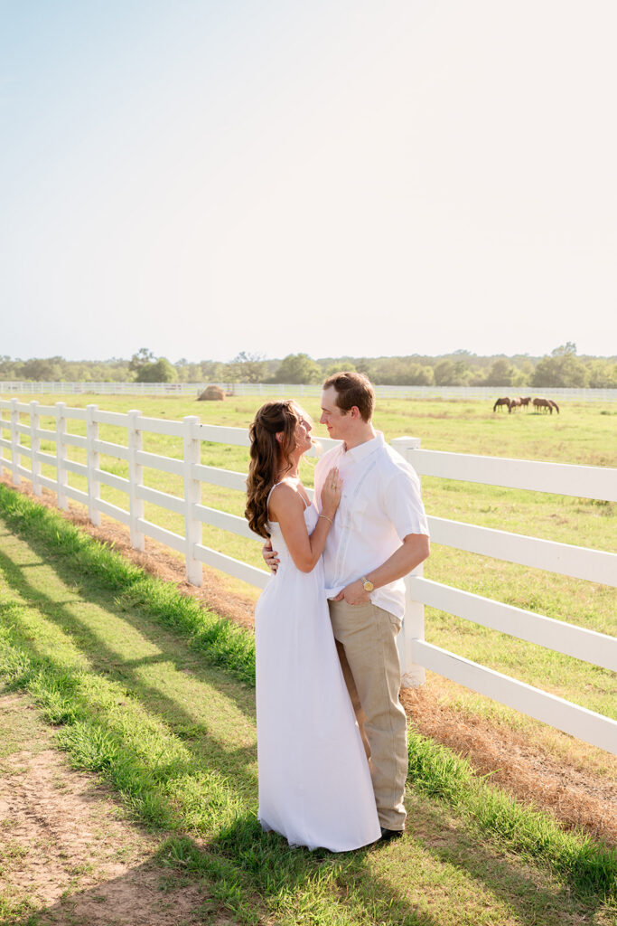 Adalyn and Miles lean against the white ranch fence laughing together, with grazing horses and a hay bale in the pasture behind them.