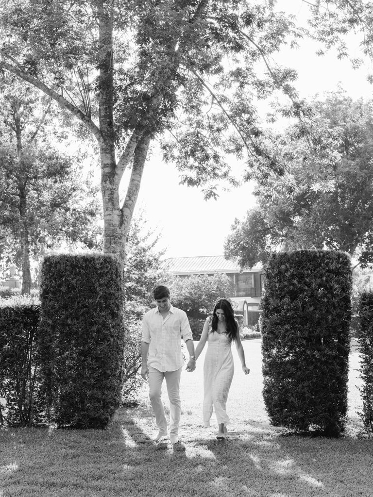 Black and white photo of the engaged couple walking hand in hand between tall hedges at Bayou Bend in Houston